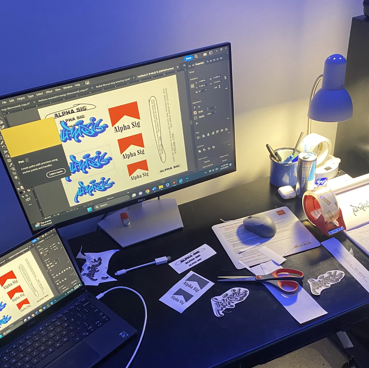 A design workstation with Alpha Sig artwork on screen and printed proofs on the desk